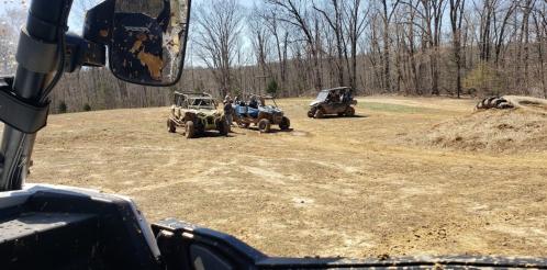 View from an off-road vehicle showing several ATVs parked on a muddy field surrounded by trees.