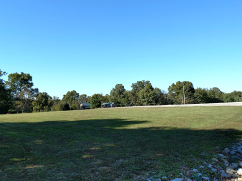 A clear blue sky over a grassy field with trees in the background and a gravel edge in the foreground.