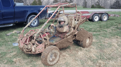 A person in a muddy go-kart sits on grass, wearing a helmet, with a truck and trailer in the background.