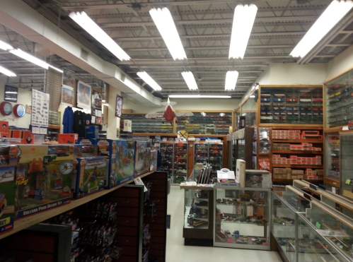 A well-stocked hobby shop interior with shelves of model kits, collectibles, and display cases. Bright overhead lighting.