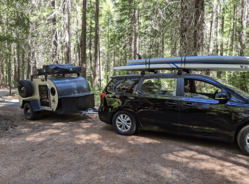 A black minivan towing a small camper trailer in a forested area, with kayaks on the roof.