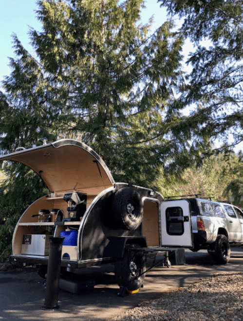 A compact camper trailer with an open kitchen area parked beside a truck, surrounded by tall green trees.