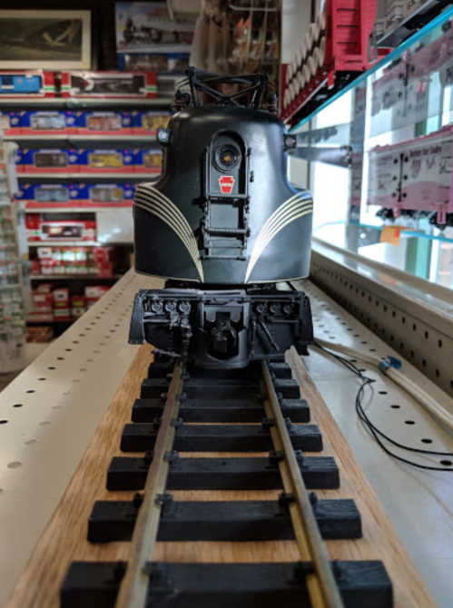 A black model train engine on a track, displayed in a store with colorful model kits in the background.