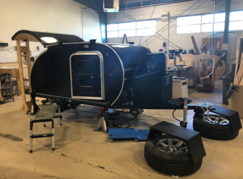 A black teardrop trailer in a workshop, with tools and equipment visible in the background.