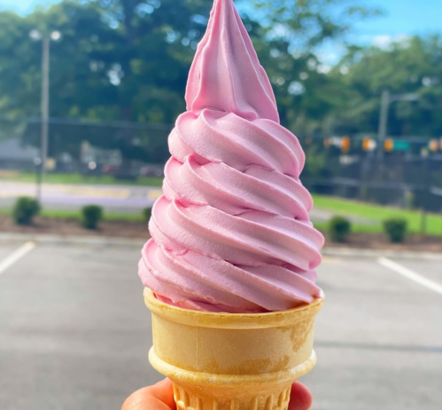 A hand holding a large swirl of pink soft serve ice cream in a cone, with a blurred outdoor background.