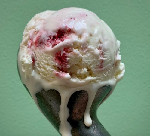 A close-up of a melting scoop of ice cream with swirls of pink and white, resting on a gray cone against a green background.
