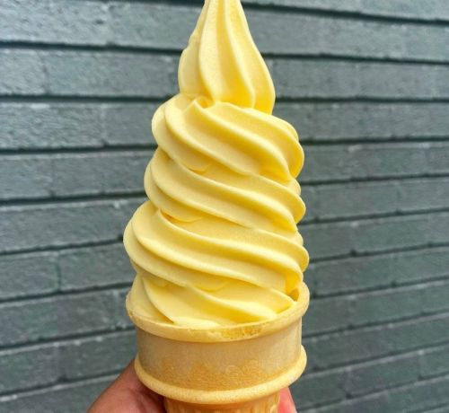 A hand holding a tall, swirled yellow soft serve ice cream cone against a gray brick wall.