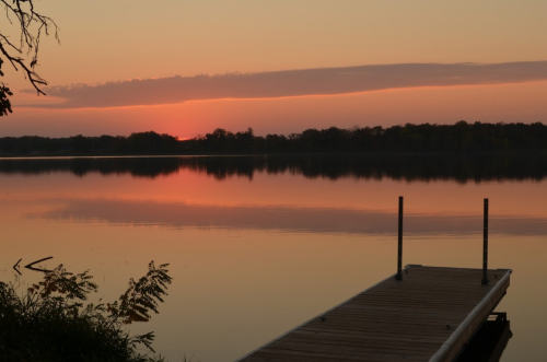 A serene lake at sunset, with a wooden dock extending into calm waters reflecting pink and orange hues.