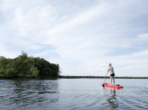 A person paddleboarding on a calm lake, surrounded by trees and under a partly cloudy sky.
