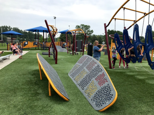A colorful playground with swings and climbing structures, surrounded by green grass and shaded areas. Families are playing.