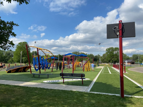 A colorful playground with slides and climbing structures, a basketball hoop, and green grass under a blue sky.