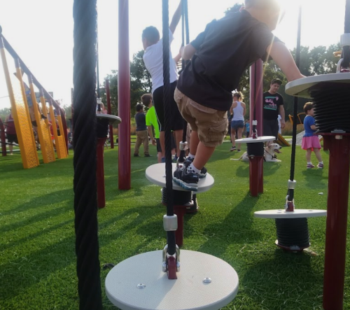 Children play on a climbing structure in a vibrant playground, surrounded by other kids and green grass.