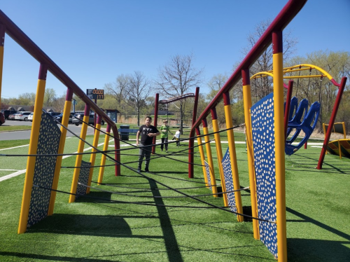A child stands in a playground with colorful equipment and a web of ropes, under a clear blue sky.