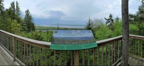 A scenic overlook with a sign, surrounded by trees, showcasing a view of a lake and distant landscape under cloudy skies.