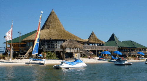 Tropical beach scene with thatched-roof buildings, jet skis, and blue umbrellas under a clear sky.