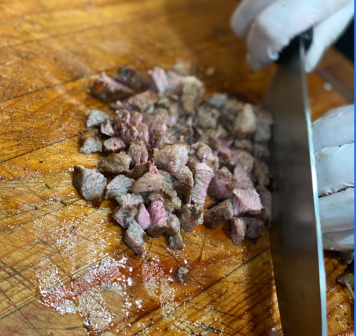 A close-up of finely chopped meat on a wooden cutting board, with a hand holding a knife in the background.