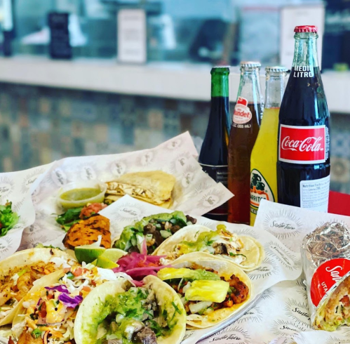 A colorful spread of tacos with various toppings, accompanied by drinks and lime wedges on a table.