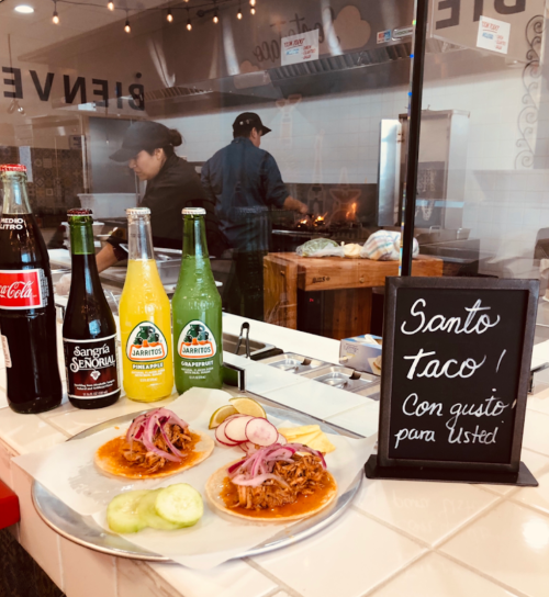 Two tacos on a plate with radishes and cucumbers, alongside drinks and a sign that reads "Santo taco! Con gusto para Usted."