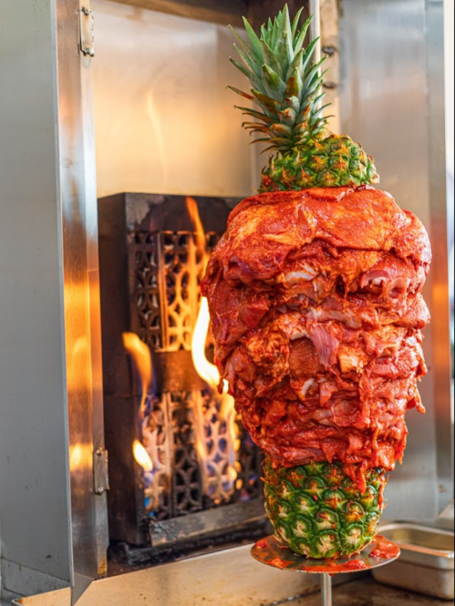 A vertical skewer of marinated meat topped with a whole pineapple, cooking over an open flame.