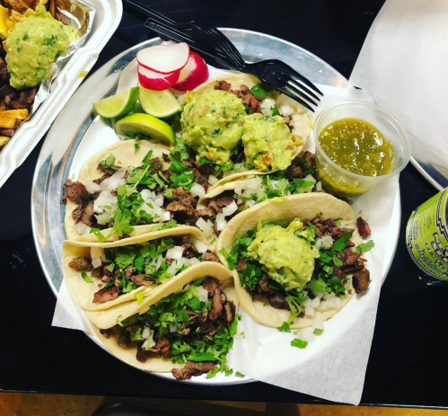 A plate of tacos filled with meat, onions, and cilantro, topped with guacamole, served with lime, radishes, and salsa.