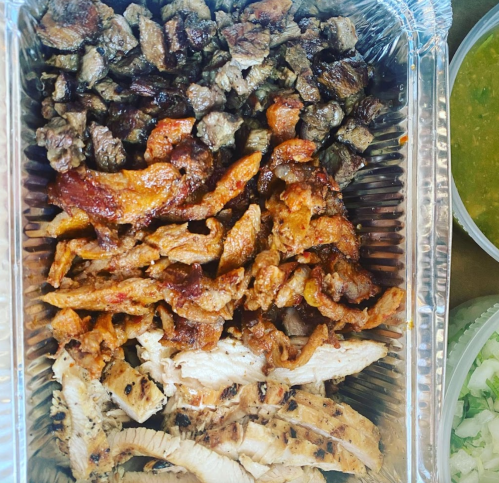 A metal tray filled with assorted grilled meats, including beef, chicken, and pork, alongside a bowl of green sauce.