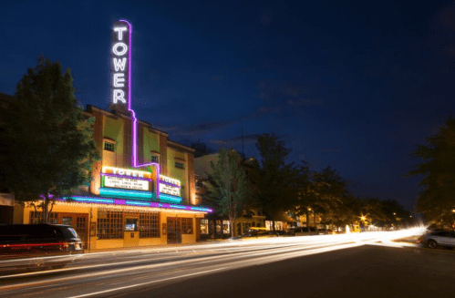 A vintage theater with a neon "TOWER" sign, illuminated at night, surrounded by trees and light trails from passing cars.