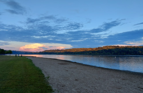 A serene lakeside view at sunset, with a sandy beach and lush green hills in the background.