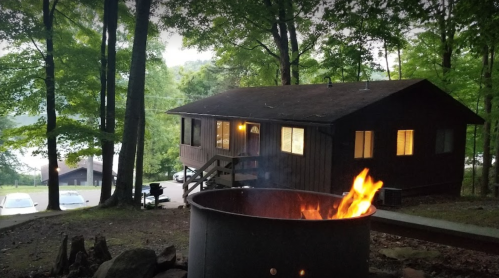 A cozy cabin in the woods with a fire pit in the foreground, surrounded by trees and a serene atmosphere.