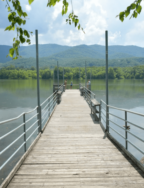 A wooden pier extends over calm water, surrounded by green mountains and a cloudy sky. Two people stand at the end.