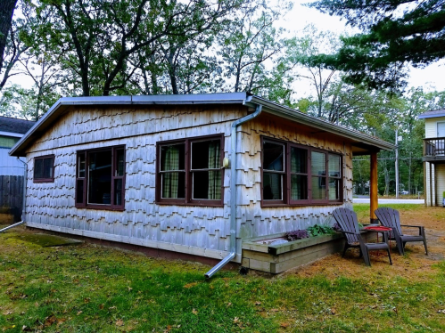 A wooden cabin with a shingled exterior, surrounded by trees and featuring a small patio with two chairs.