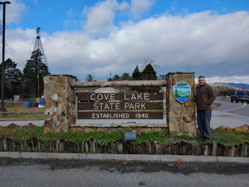 Sign for Cove Lake State Park, established in 1940, with a person standing beside it and a cloudy sky in the background.