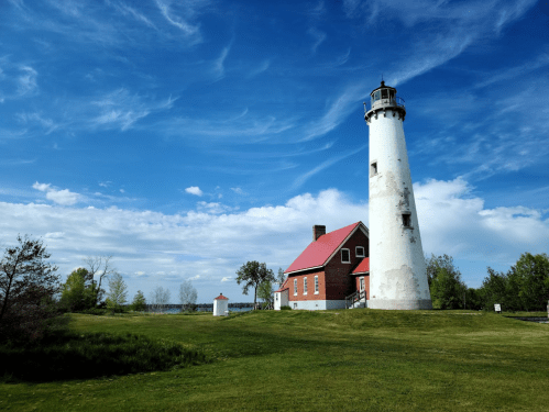 A tall white lighthouse with a red roof stands on green grass under a blue sky with wispy clouds.