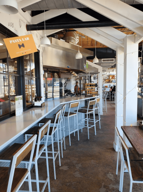 Interior of a modern restaurant featuring a bar with white stools and an open kitchen area. Bright and airy atmosphere.