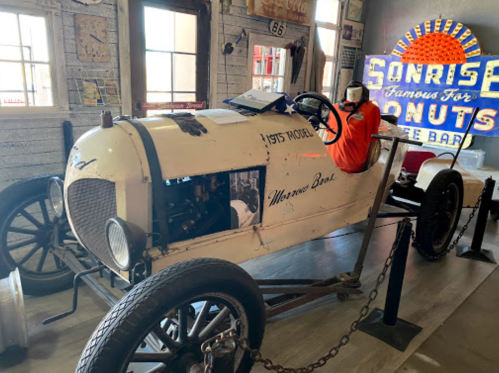 A vintage race car displayed indoors, next to a colorful sign for Sunrise Donuts.
