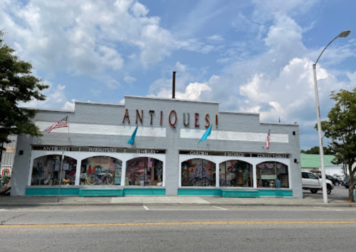 A vintage shop named "Antiques" with large windows displaying various items, set against a cloudy sky.