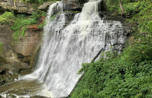 A cascading waterfall surrounded by lush greenery and rocky terrain. Water flows down in a misty spray.