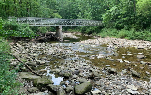 A metal bridge spans a shallow creek surrounded by lush green trees and rocky banks.