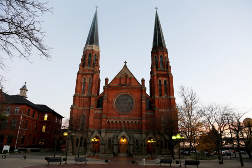 Historic red brick church with two tall spires, set against a twilight sky, surrounded by trees and benches.