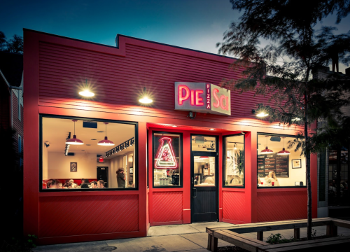 A vibrant red restaurant exterior with a neon sign reading "Pie" and "Sol," featuring outdoor seating and warm lighting.