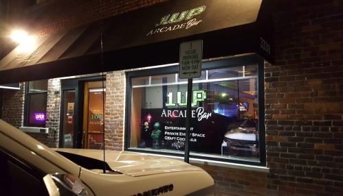 Exterior view of 1UP Arcade Bar at night, featuring a lit sign and large windows showcasing the interior.