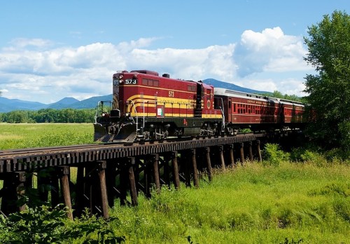 A red and yellow train crosses a wooden trestle bridge surrounded by green fields and mountains under a blue sky.