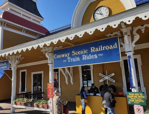 Exterior of the Conway Scenic Railroad station, featuring a sign for train rides and people at the ticket counter.