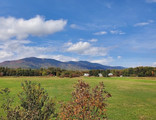 A scenic view of mountains under a blue sky, with green fields and autumn foliage in the foreground.