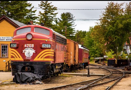 A vintage red and yellow train engine parked beside a wooden station, surrounded by trees and railway tracks.