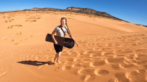A girl with braids walks on sand dunes, holding a snowboard, under a clear blue sky.