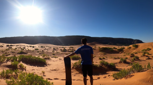 A person stands in a desert, holding a snowboard, with sun shining brightly and dunes in the background.