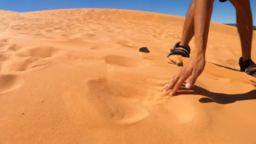 A close-up of a hand touching the smooth, orange sand of a desert dune under a clear blue sky.