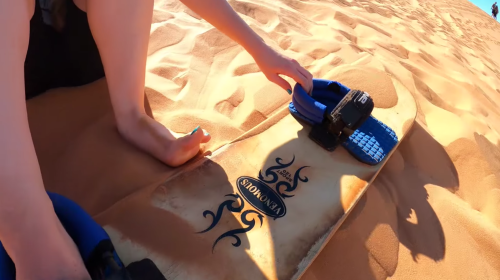 A person prepares a sandboard on a sandy dune, with blue bindings and a decorative design on the board.