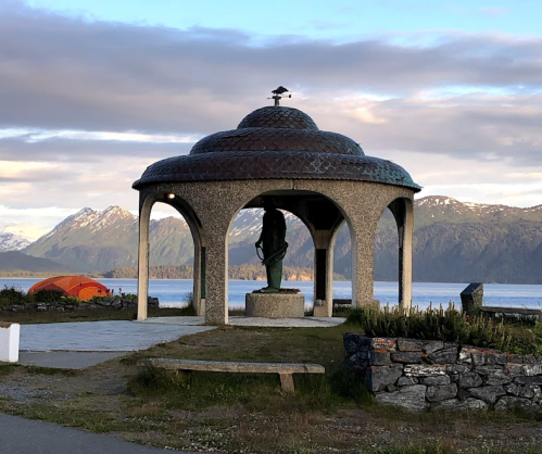 A stone gazebo with a statue inside, overlooking a lake and mountains, with a tent nearby.