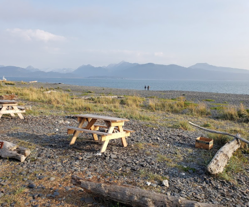 A serene beach scene with picnic tables, rocky shore, and distant mountains under a clear sky. Two people walk along the water.